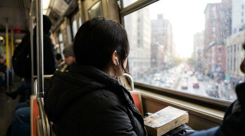 Over the shoulder of a woman on a morning commuter train listening with white earbuds, a paperback on her lap, the city passing outside the window