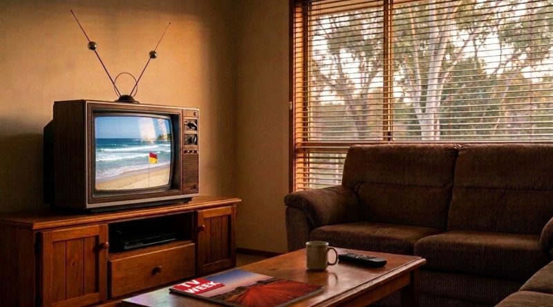 A 1990s Australian lounge room with a CRT television showing a surf beach and a red and yellow flag, a corduroy couch, and a TV Week magazine on the coffee table