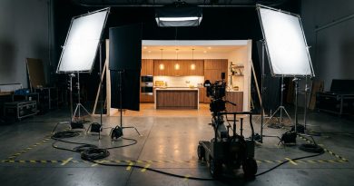 An empty television commercial set on a soundstage: a two-walled modern kitchen lit by softboxes, with light stands, cables taped to the floor and a cinema camera on a dolly