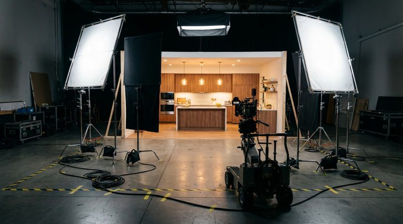 An empty television commercial set on a soundstage: a two-walled modern kitchen lit by softboxes, with light stands, cables taped to the floor and a cinema camera on a dolly