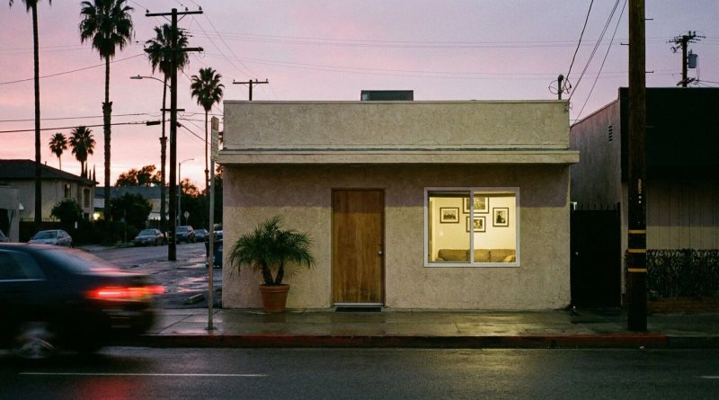A small beige stucco recording studio on a North Hollywood street at dusk, its lobby light still on behind the window, palm trees and power lines against a pink sky. © The Voice Realm