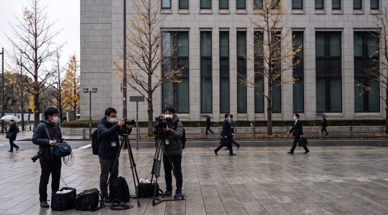 Fotógrafos de prensa y camarógrafos esperan con trípodes en una acera mojada frente a un edificio gubernamental de piedra gris en el distrito de Kasumigaseki, Tokio. © The Voice Realm