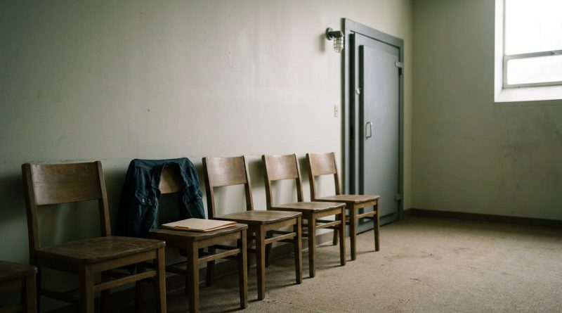 An empty waiting room outside a recording session: a row of wooden chairs against a pale wall, a jacket and a paper folder left on one seat, a closed grey studio door at the end