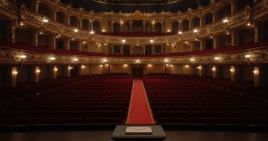 An empty awards theatre before the ceremony, seen from the stage: rows of red velvet seats, a red centre aisle, gilded balconies lit by house lights, and a closed envelope on the lectern