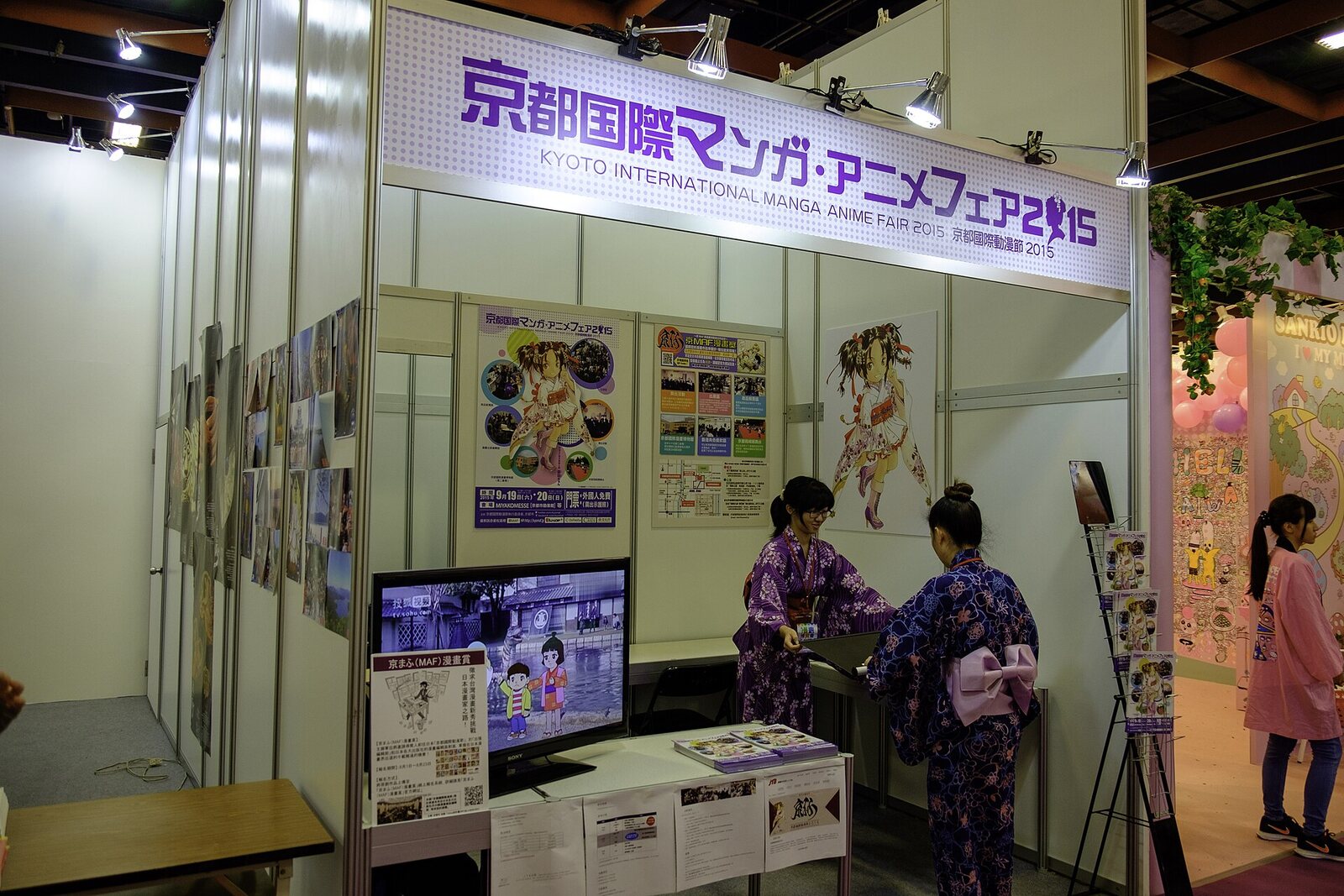 Un stand de la Feria Internacional de Manga y Anime de Kioto en 2015, con el cartel morado de la feria, pósters de anime y dos empleadas en yukata junto a una pantalla