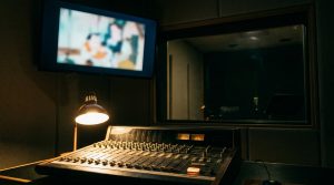 The director's side of a dubbing session: a mixing console under a desk lamp, a monitor showing a blurred frame of animation, and a dark empty booth through the glass