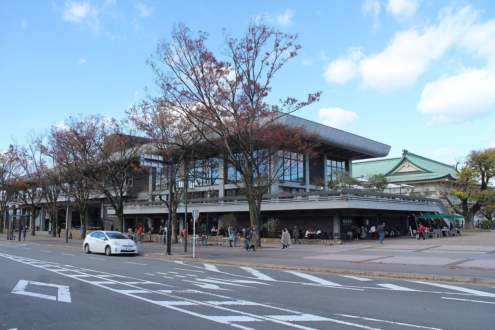 El ROHM Theatre Kyoto, un edificio moderno y alargado de techo bajo inclinado detrás de árboles otoñales y una calle ancha bajo un cielo azul
