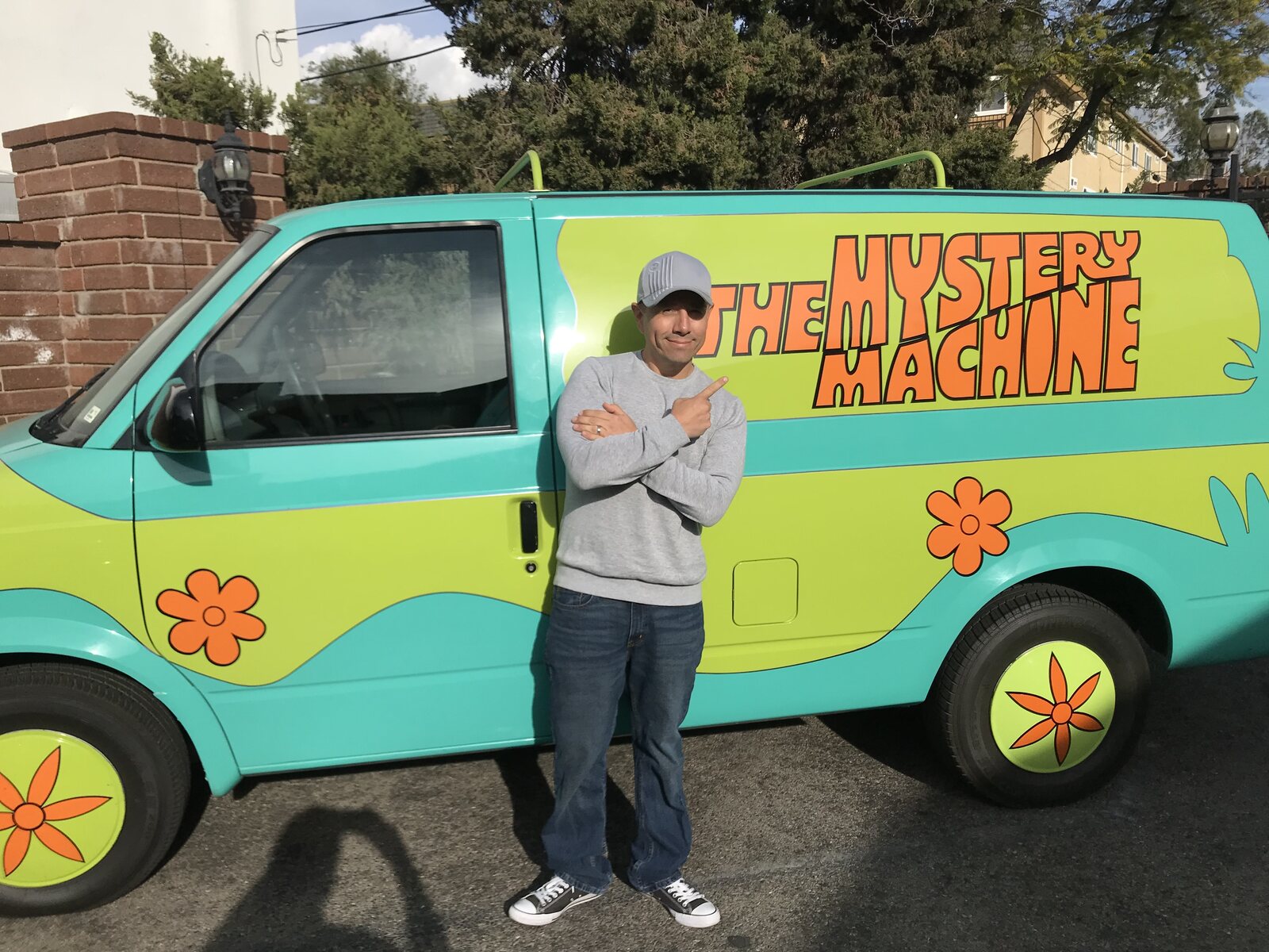 A man in a grey hoodie stands beside a van painted as the Mystery Machine, with flowers and the words The Mystery Machine on its side. Image: Salami Studios
