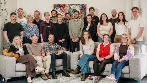 The Abe's Audio team, about twenty people, pose on and around two grey sofas in front of a colourful floral painting in their office. Image: Abe's Audio
