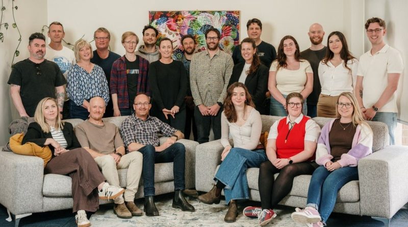 The Abe's Audio team, about twenty people, pose on and around two grey sofas in front of a colourful floral painting in their office. Image: Abe's Audio