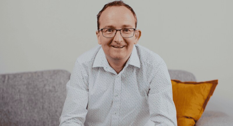 Abe Udy, founder of Abe's Audio, smiling on a grey sofa in a white patterned shirt in a 2024 company photo. Image: Abe's Audio