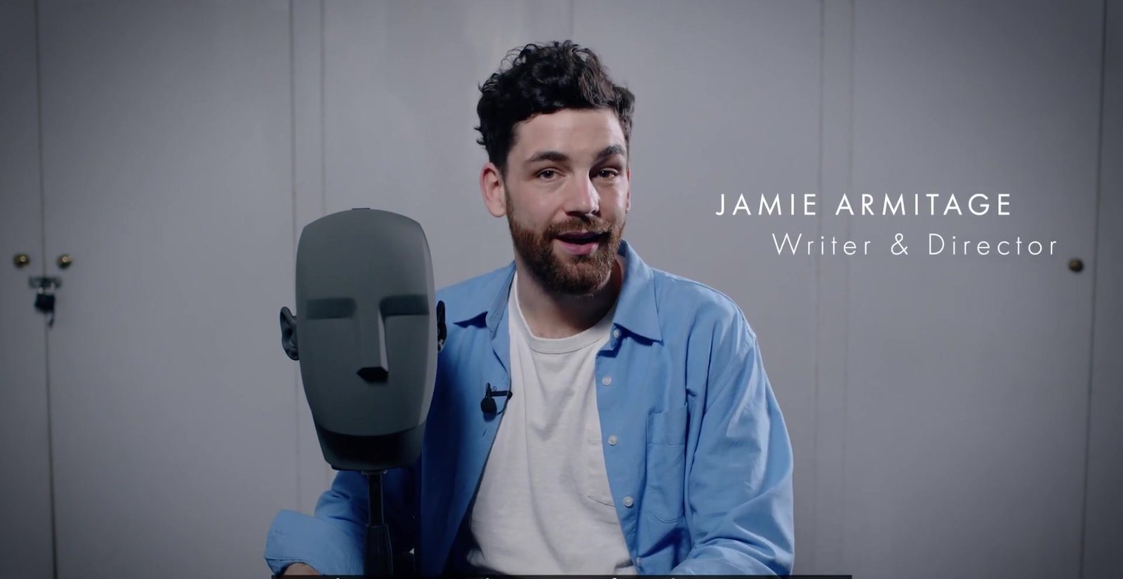 Writer and director Jamie Armitage, in a blue shirt, sits beside the grey binaural dummy head used in A Ghost in Your Ear. Image: Hampstead Theatre