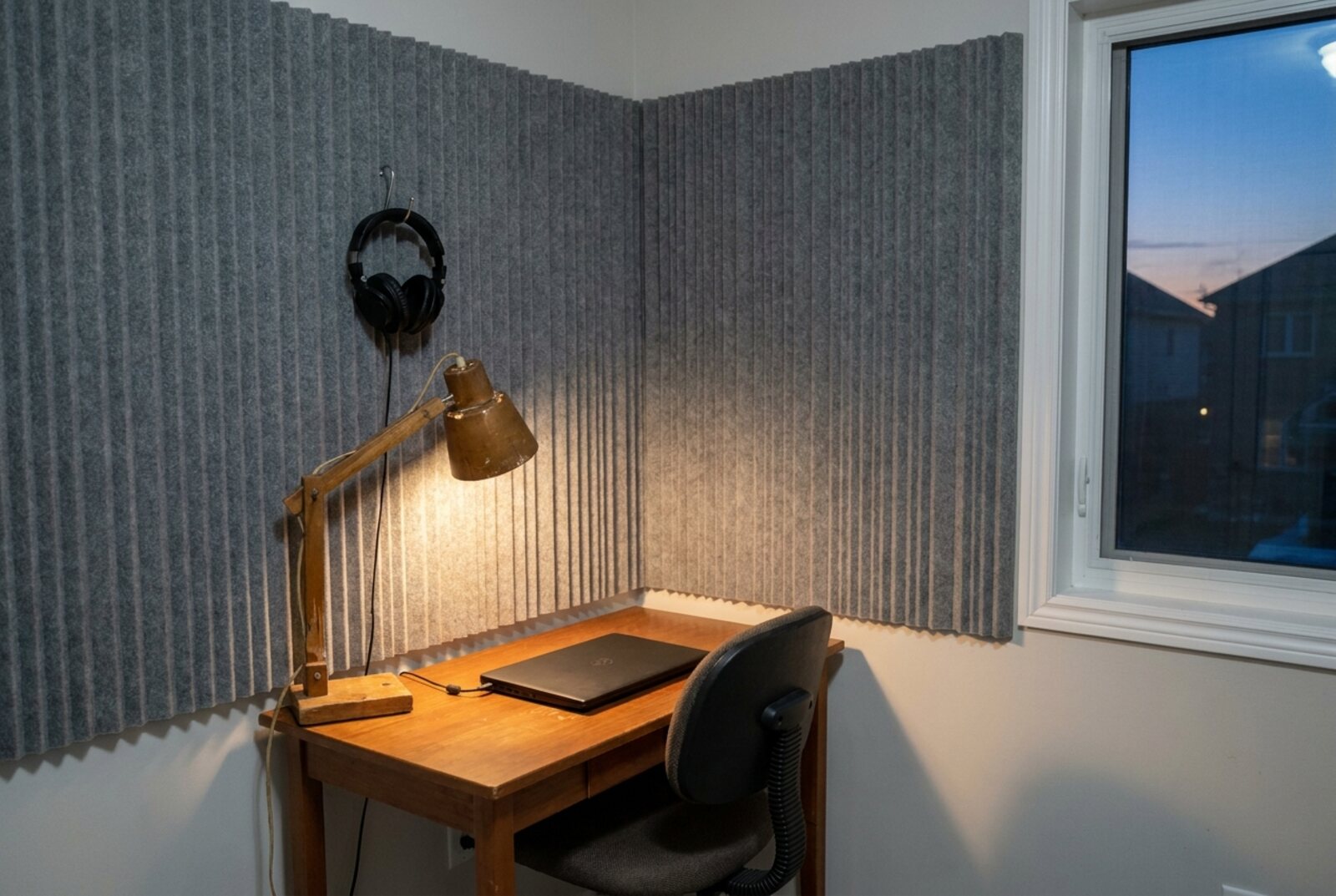 Una cabina de locución casera vacía al atardecer, con audífonos colgados en la espuma y una laptop cerrada sobre el escritorio. © Voice Over Herald