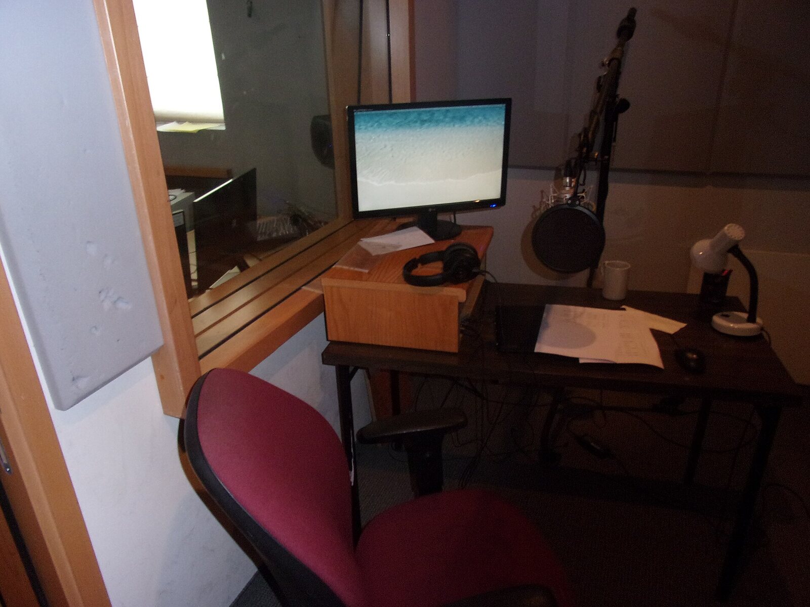 Una cabina de doblaje vacía: un monitor sobre un soporte de madera, audífonos, un guion acostado sobre la mesa, una lámpara de escritorio y un micrófono de condensador detrás de un filtro antipop. Foto: Tamirsofer, Wikimedia Commons, CC BY-SA 4.0