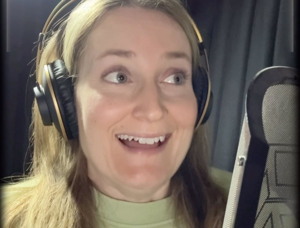 Emily McGonagle in her home recording booth wearing headphones beside a studio microphone, from one of her behind the scenes videos. Image: Emily McGonagle