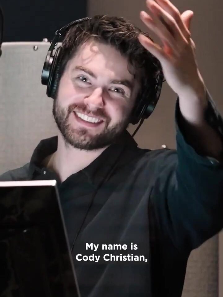 Cody Christian, the English voice of Cloud Strife, smiles and waves in a recording booth, captioned My name is Cody Christian, a frame from Square Enix's Behind the Booth video. Image: Square Enix