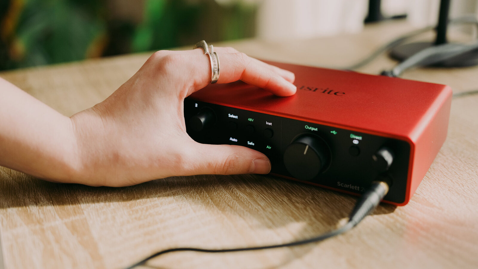 A hand resting on a red Focusrite Scarlett 2i2 fourth generation audio interface on a wooden desk, with a cable plugged into the front. Image: Focusrite