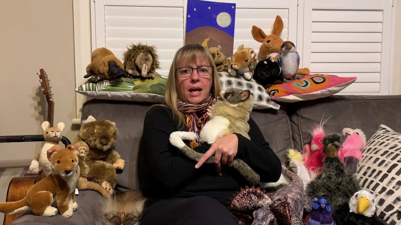 La autora Helen Milroy, sentada en un sofá gris rodeada de peluches de animales australianos y con una zarigüeya de peluche, lee en voz alta en un video de Fremantle Press. Imagen: Fremantle Press