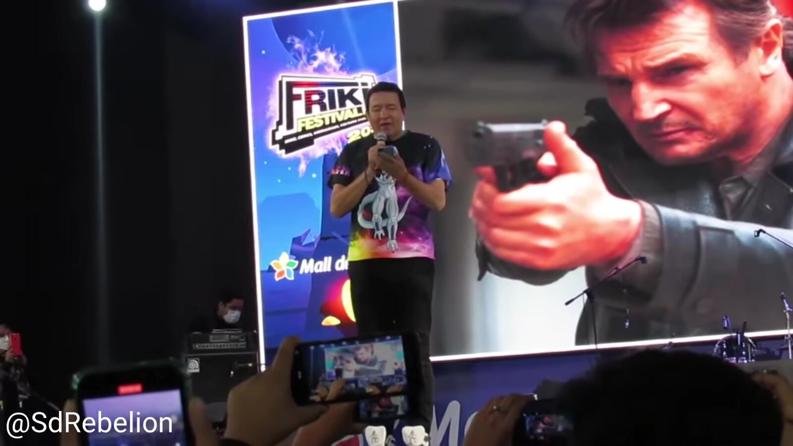 Gerardo Reyero on stage at Friki Festival 2022, microphone in hand, doing Liam Neeson's voice in front of a giant still of Neeson aiming a pistol in Taken. Image: Sombras de Rebelión