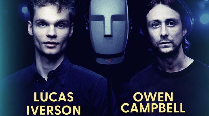 Official cast image for A Ghost in Your Ear: Lucas Iverson and Owen Campbell on either side of a grey binaural dummy head microphone, under blue stage light. Image: Audible Theater