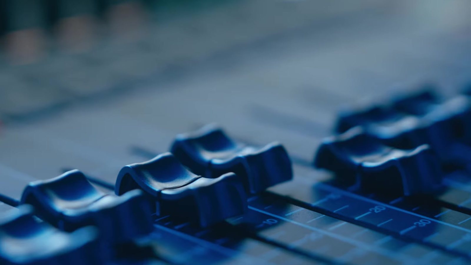 Close view of faders on a sound desk in blue light, from the trailer for A Ghost in Your Ear. Image: Audible Theater