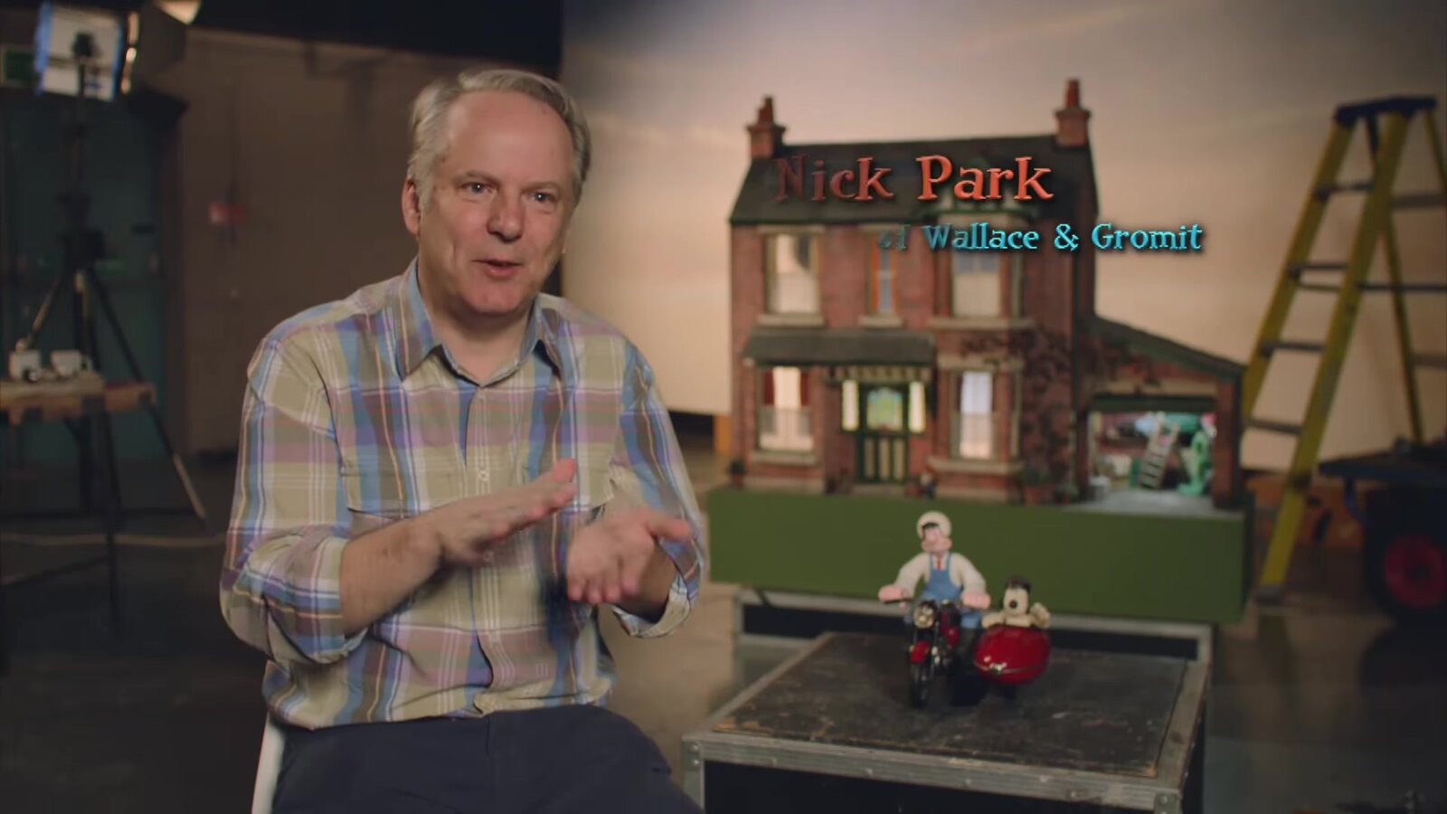 Nick Park, con camisa a cuadros, habla junto a una maqueta de la casa de Wallace y Gromit con la pareja en una moto con sidecar al frente, con su nombre en pantalla, un fotograma de The Making of A Grand Day Out en el canal oficial de Wallace y Gromit. Imagen: Aardman