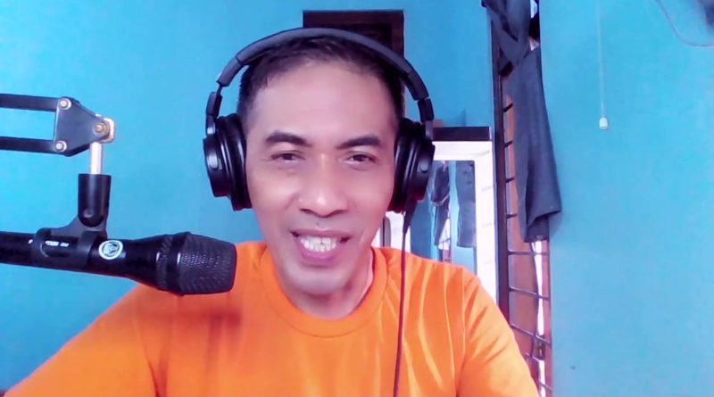 Toto Warsito sonríe frente a un micrófono, con audífonos y camiseta naranja, en su lección Belajar Dubbing en YouTube. Imagen: Toto Warsito, PAKDE TO en YouTube