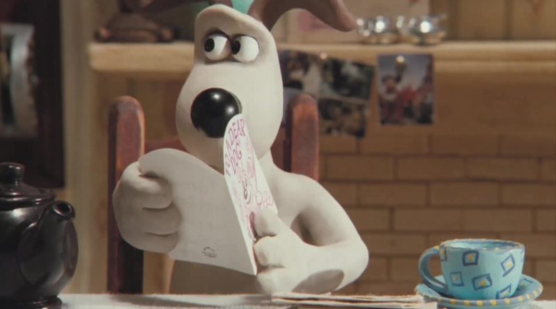 Gromit, el perro de plastilina de Wallace y Gromit, sentado a la mesa de la cocina junto a una tetera y una taza azul leyendo una tarjeta que dice To a dear dog, un fotograma del documental de Aardman A Grand Night In en el canal oficial de Wallace y Gromit. Imagen: Aardman
