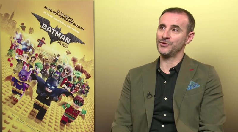 Spanish dubbing actor Claudio Serrano smiles during a video interview, seated beside a poster for The Lego Batman Movie. Image: Moobys