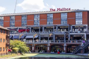 The Mailbox in Birmingham, a red brick building with a red The Mailbox sign on its roof, above canalside restaurants. Photo: The wub, CC BY-SA 4.0, via Wikimedia Commons