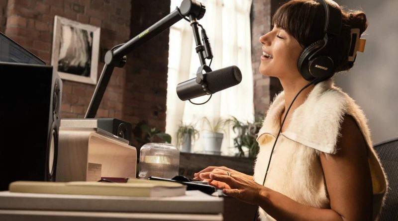 Un micrófono Shure SM7B en un brazo articulado en un estudio casero luminoso, con una mujer con audífonos que habla frente a él en su escritorio. Imagen: Shure