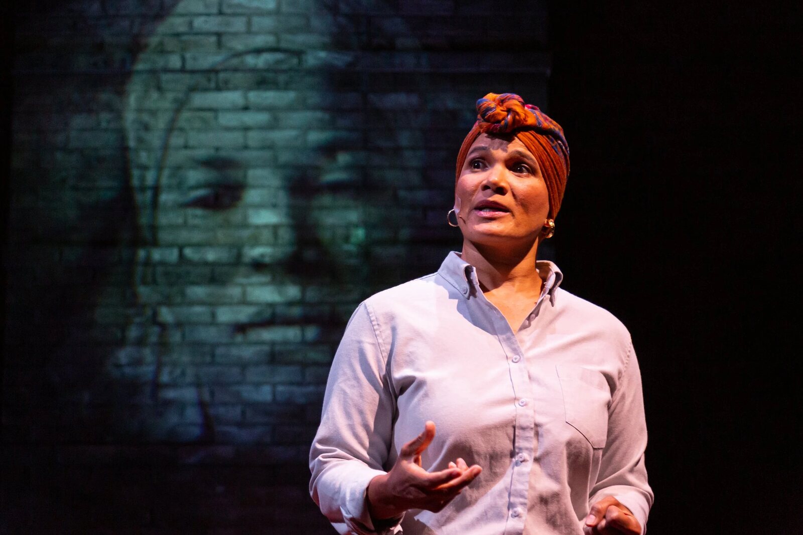 January LaVoy on stage in a headwrap and pale blue shirt, a projected face on a brick wall behind her. Image: januarylavoy.com