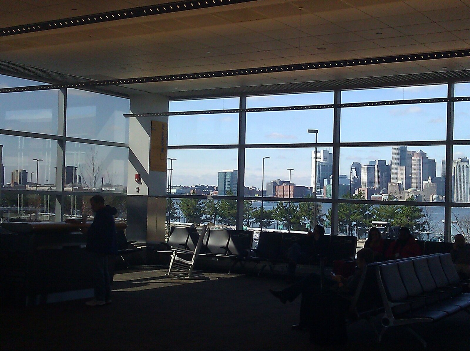 Una sala de embarque del aeropuerto Logan de Boston, con filas de asientos en sombra y el horizonte de Boston al otro lado del puerto tras grandes ventanales. Foto: Stephen Pierzchala, Wikimedia Commons, CC BY 2.0