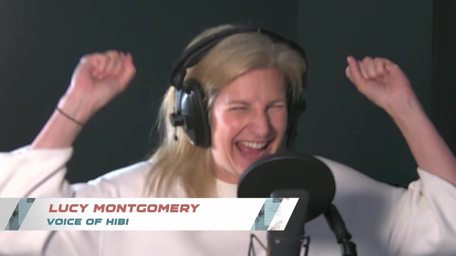 Lucy Montgomery ríe con los brazos en alto mientras graba a Hibi frente a un micrófono de estudio, con un rótulo en pantalla que dice Lucy Montgomery, Voice of Hibi. Fotograma del video Sounds of the Galactic League de Fuse Games. Imagen: Secret Mode y Fuse Games