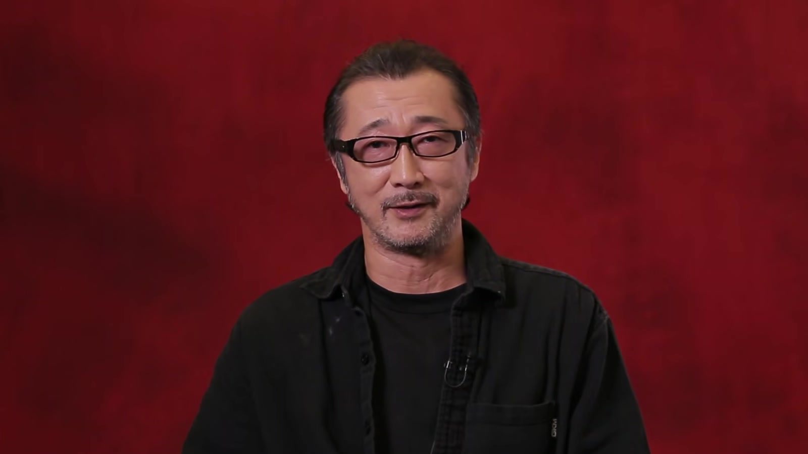 Akio Otsuka sonríe durante una entrevista frente a un fondo rojo. Imagen: RGG Studio (SEGA)