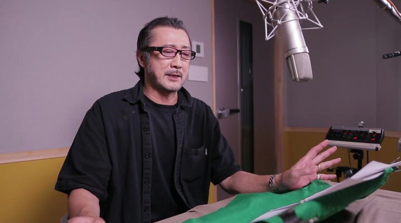 Akio Otsuka, la voz japonesa de Solid Snake, con camisa negra y lentes, habla junto a un micrófono de estudio en una cabina de grabación. Imagen: RGG Studio (SEGA)