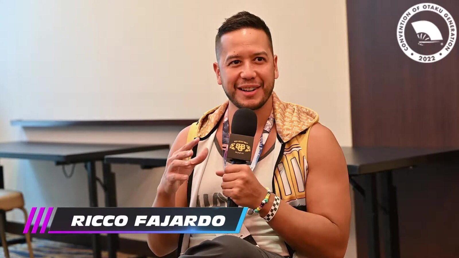 Ricco Fajardo, la voz en inglés de Grid en Overgeared, habla al micrófono durante una entrevista en Otakon 2022, con su nombre en pantalla. Imagen: Big Gold Belt Media