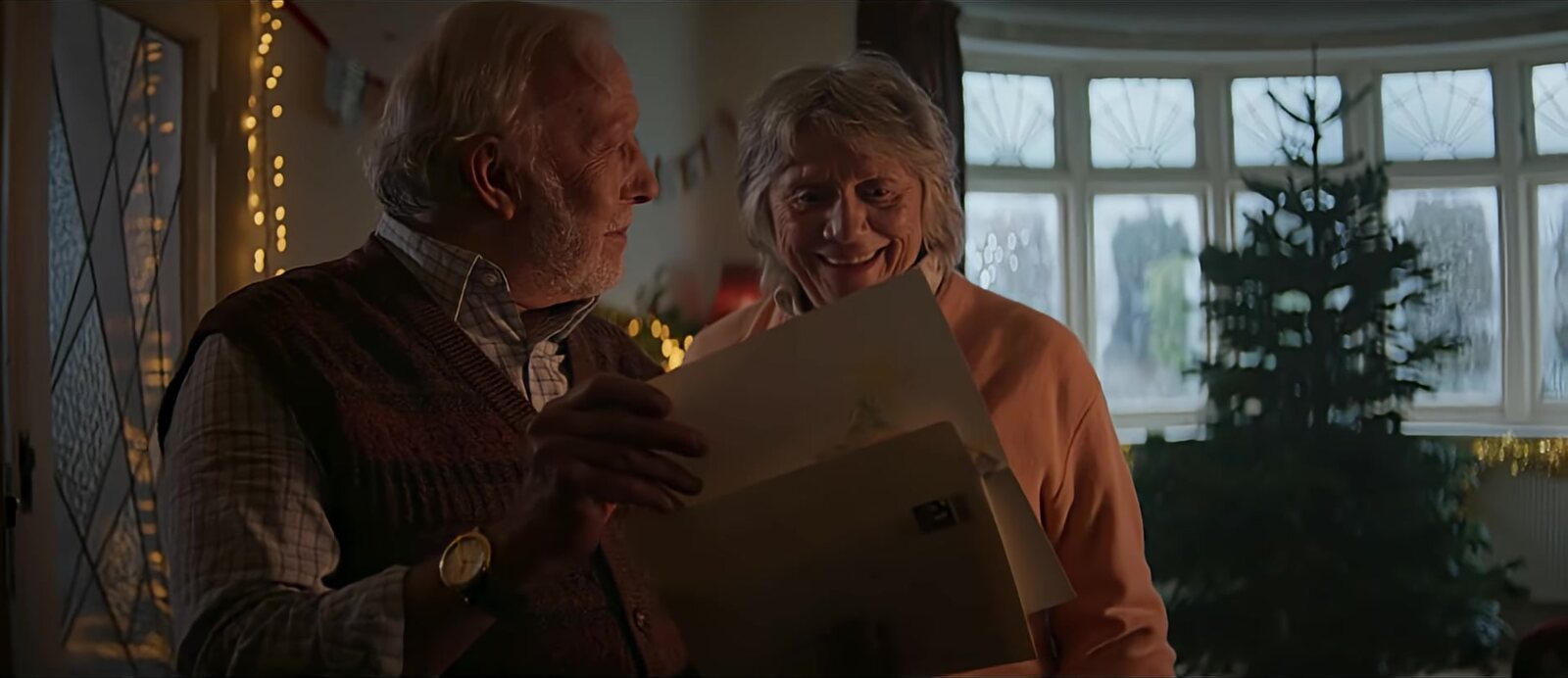 Una pareja mayor sonríe mientras lee una tarjeta navideña junto a un árbol, escena del anuncio navideño 2025 de Royal Mail. Imagen: Royal Mail