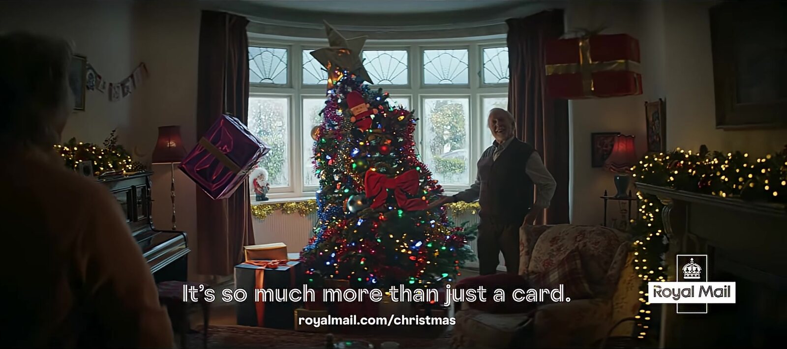 A grey-haired man stands beside a brightly lit Christmas tree in a living room under the words It's so much more than just a card and the Royal Mail logo, the closing frame of Royal Mail's 2025 Christmas ad. Image: Royal Mail