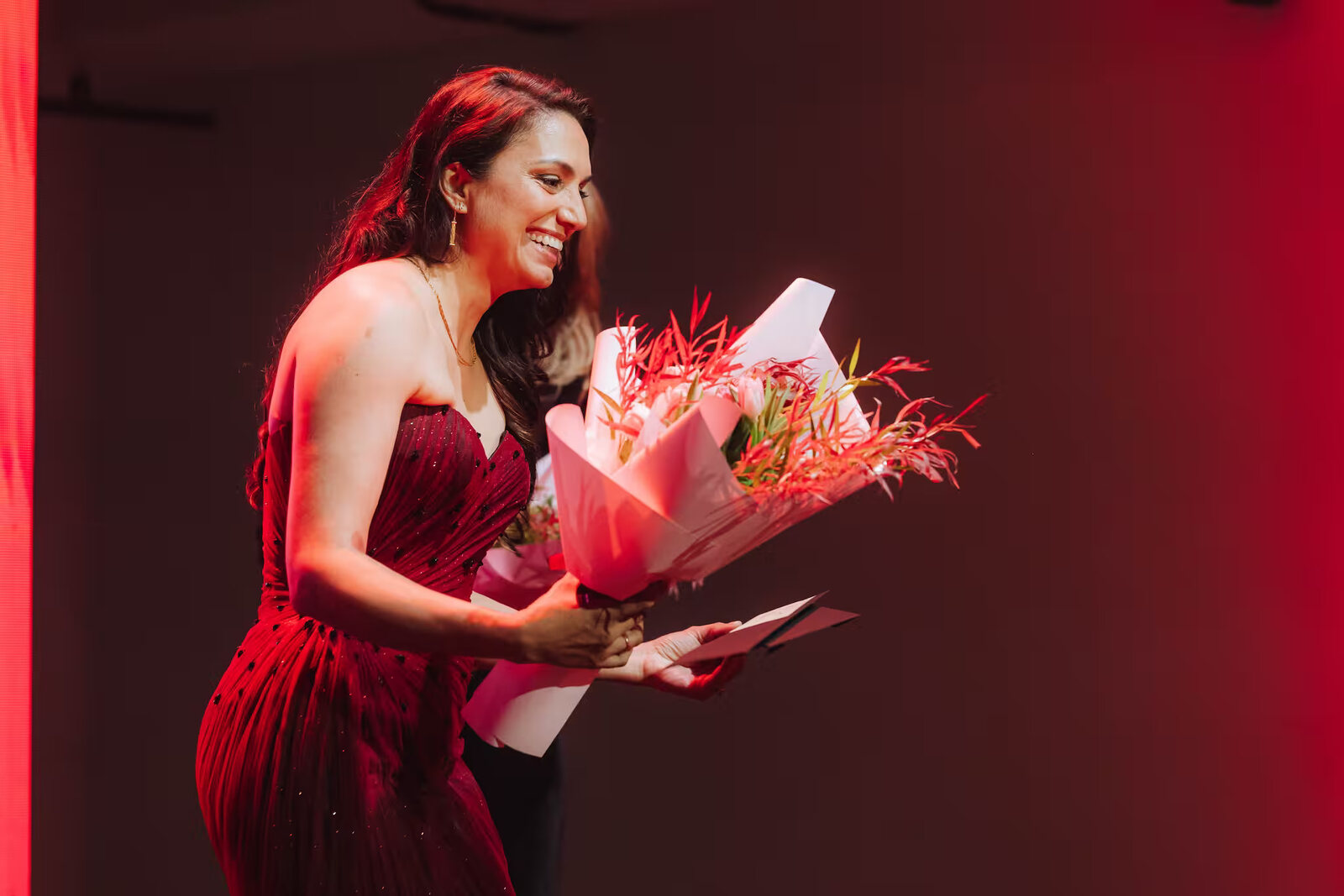 La fundadora de SONARR, Pariya Taherzadeh, sonríe en el escenario con un vestido rojo y un ramo de flores, en la gala de los Premios SONARR en Sídney. Imagen: SONARR Awards