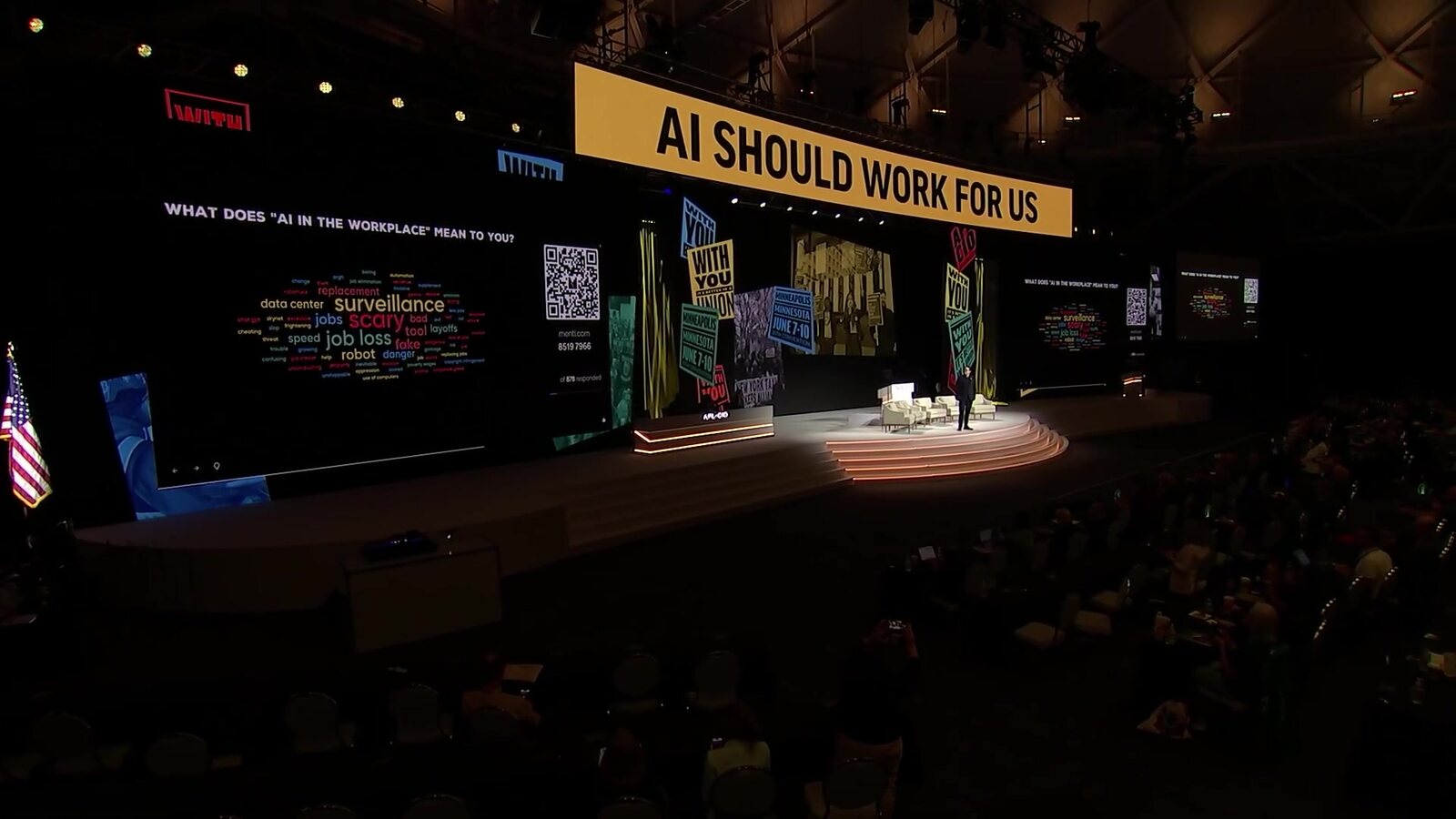 Vista amplia de un escenario bajo un cartel que dice AI Should Work For Us, con una nube de palabras sobre la IA en el trabajo en la pantalla, durante la charla de Duncan Crabtree-Ireland. Imagen: SAG-AFTRA