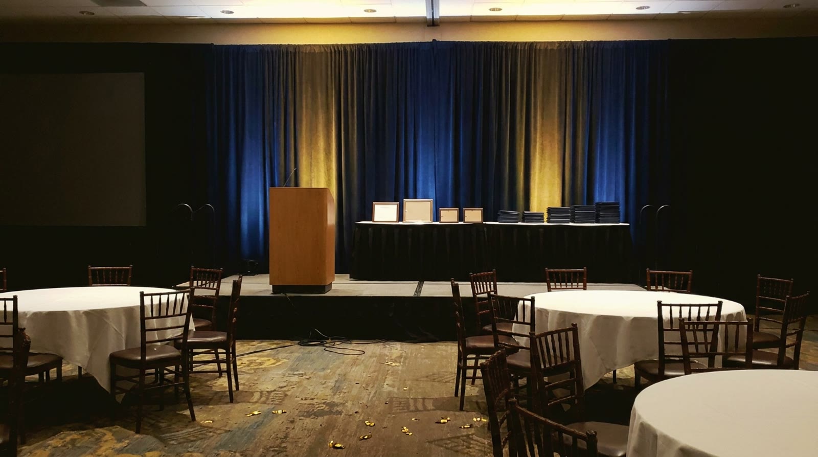 An empty hotel ballroom awards stage after a ceremony, a lectern and framed certificates on a table, gold confetti on the floor. © Voice Over Herald
