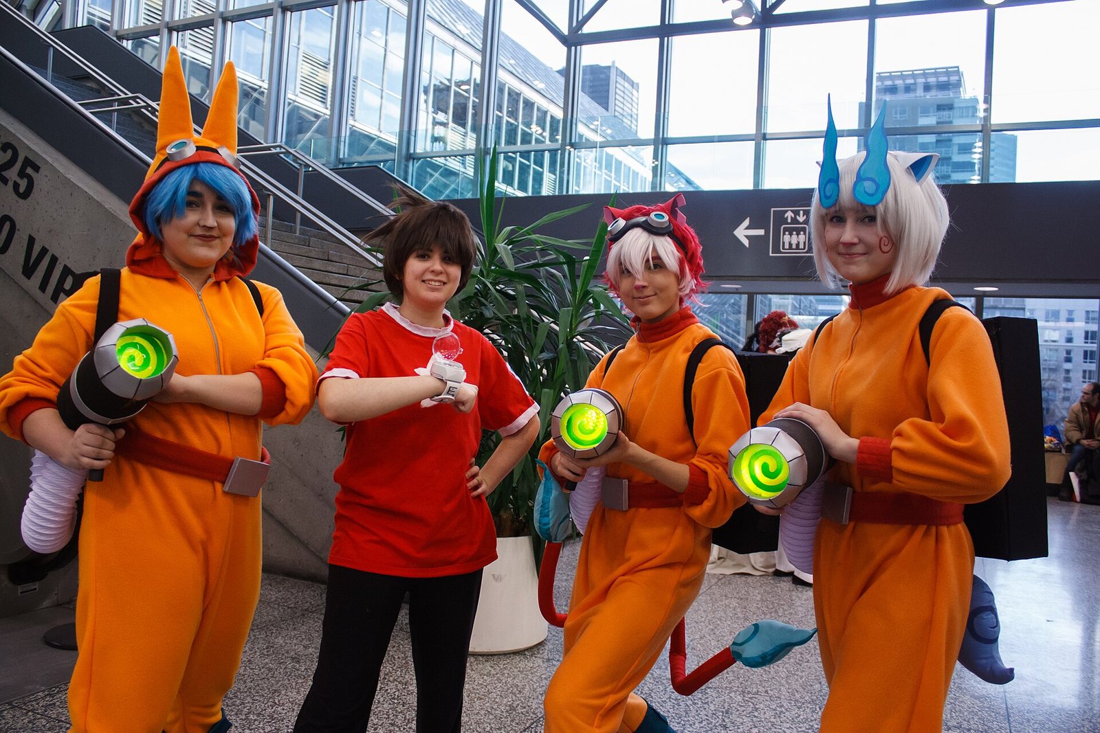 Cuatro cosplayers vestidos como personajes de Yo-kai Watch Busters, tres con trajes naranjas y lanzadores con luz verde, en el Mini-Comiccon de Montreal en 2016. Foto: Pikawil, CC BY-SA 2.0