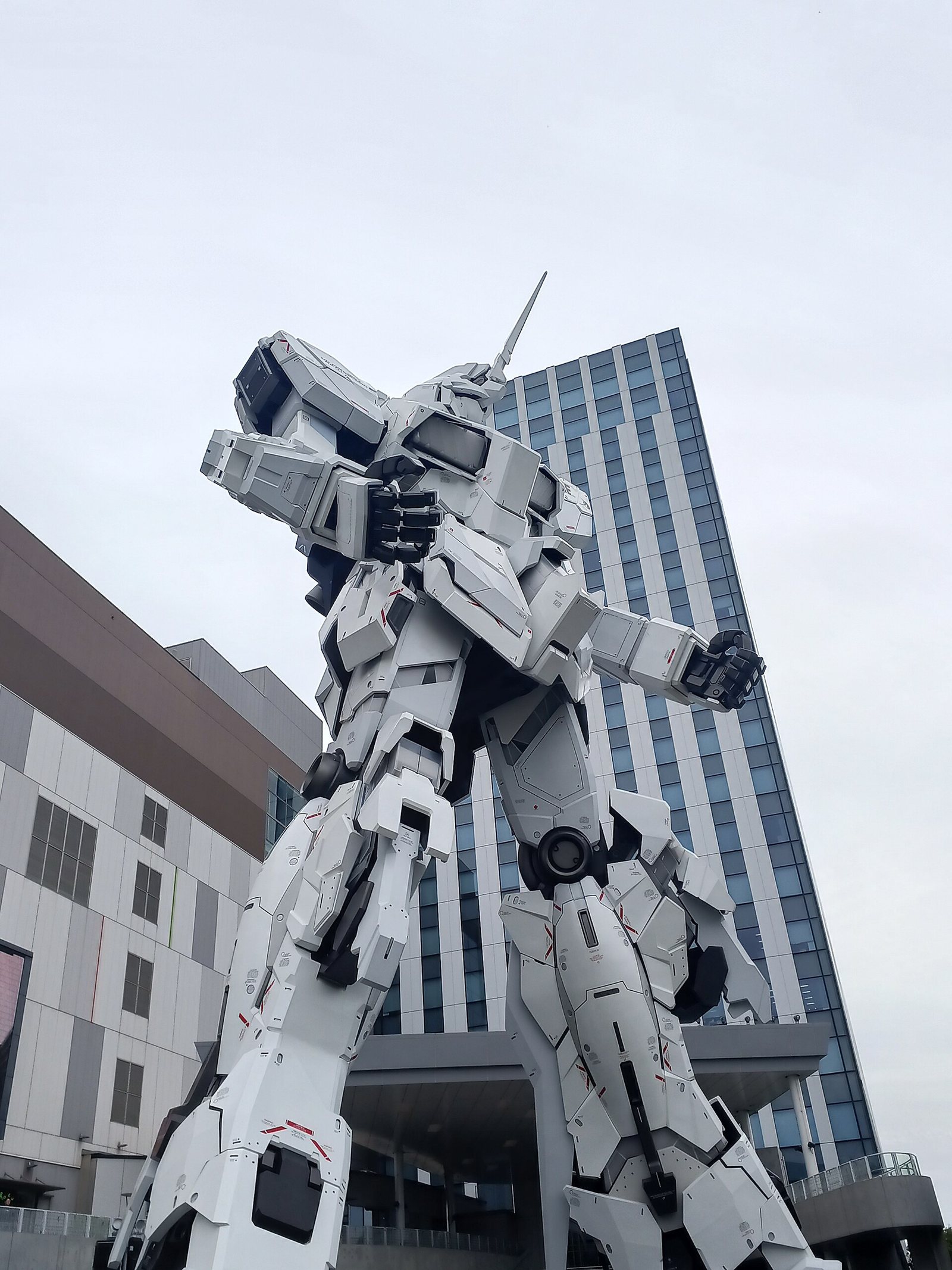 The life-size Unicorn Gundam statue in Tokyo, seen from below against a grey sky and an office tower. Photo: Pelpinosas R. Justin James, CC0