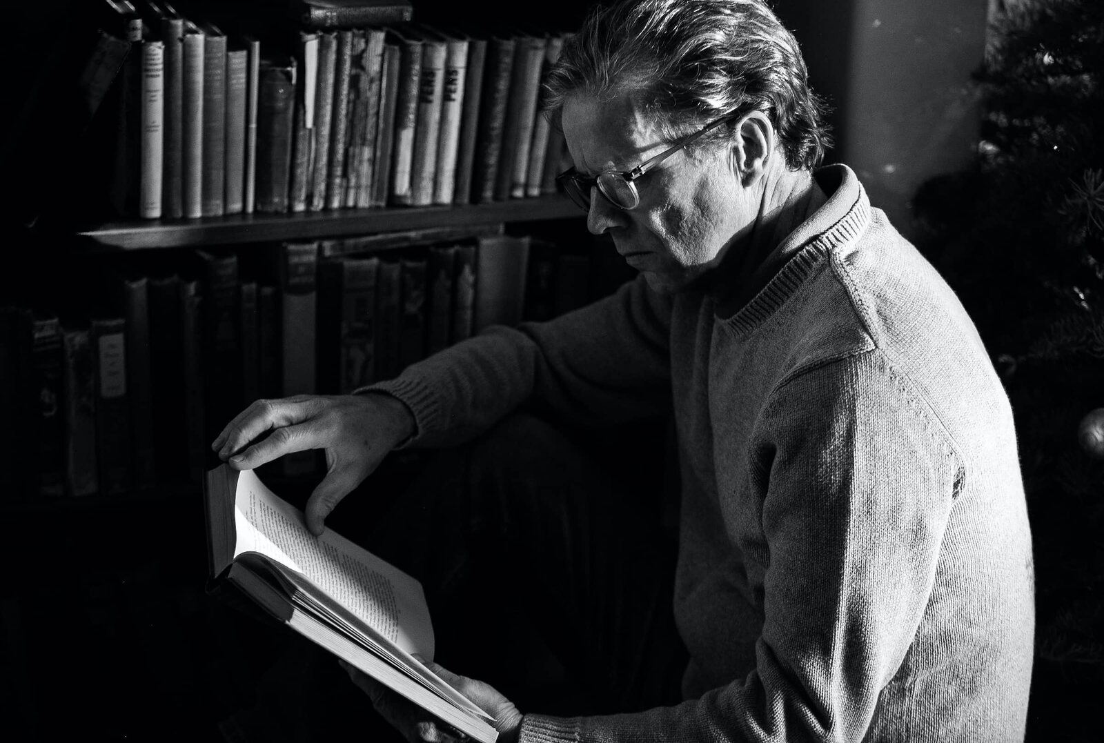 Simon Vance, con lentes y suéter, lee un libro de tapa dura junto a un estante de libros antiguos, en blanco y negro. Imagen: simonvance.com